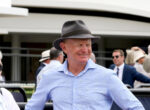 Smiling man in a light blue shirt wearing a gray fedora at an outdoor event, with others in the background.
