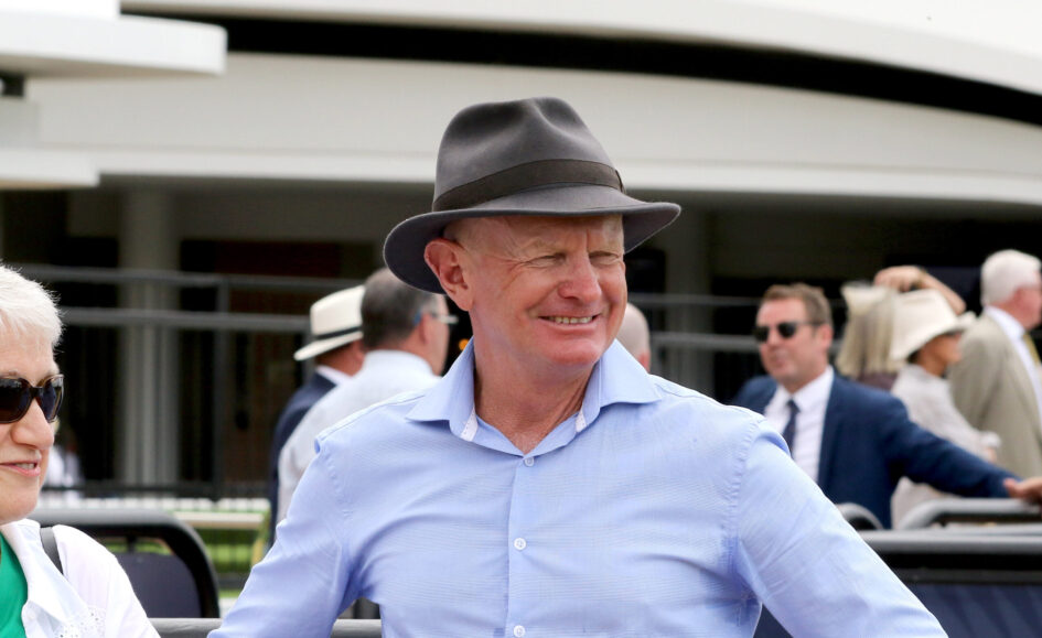 Smiling man in a light blue shirt wearing a gray fedora at an outdoor event, with others in the background.