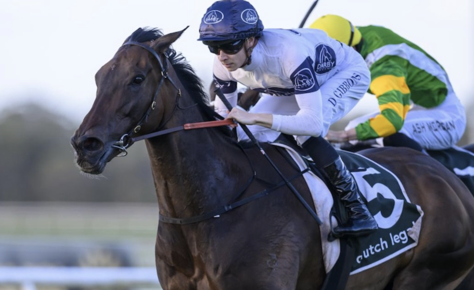 Jockey in blue helmet and white silks leans forward on a dark brown horse during a race, with a green-and-yellow rider behind in a blurred background.