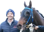 Smiling man in a blue quilted jacket and beanie standing next to a brown horse wearing a blue bridle and reins visible on its head.