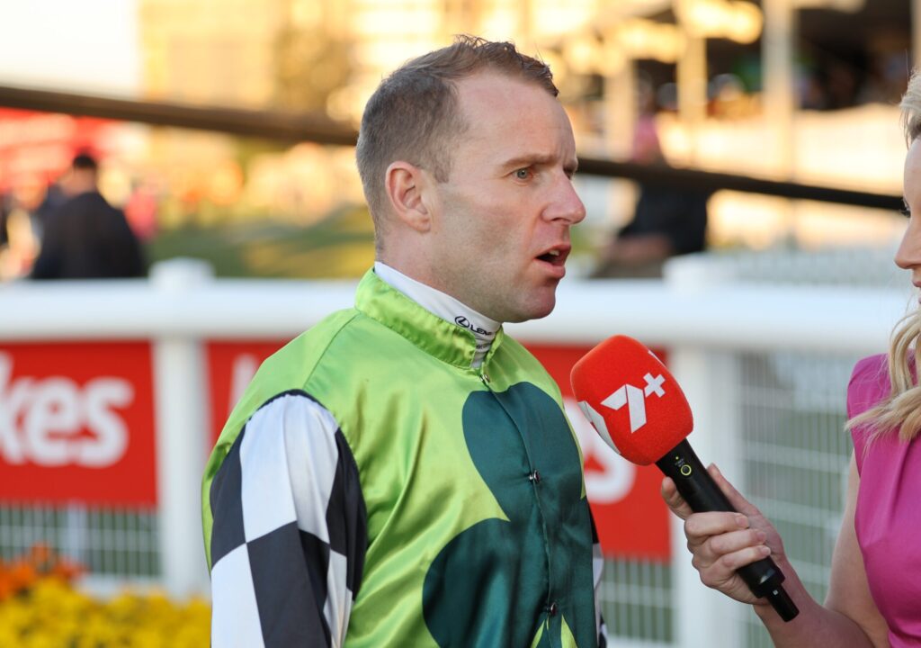 Man in a green racing suit being interviewed by a reporter with a red microphone outdoors on a sunny day.
