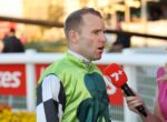 Man in a green racing suit being interviewed by a reporter with a red microphone outdoors on a sunny day.