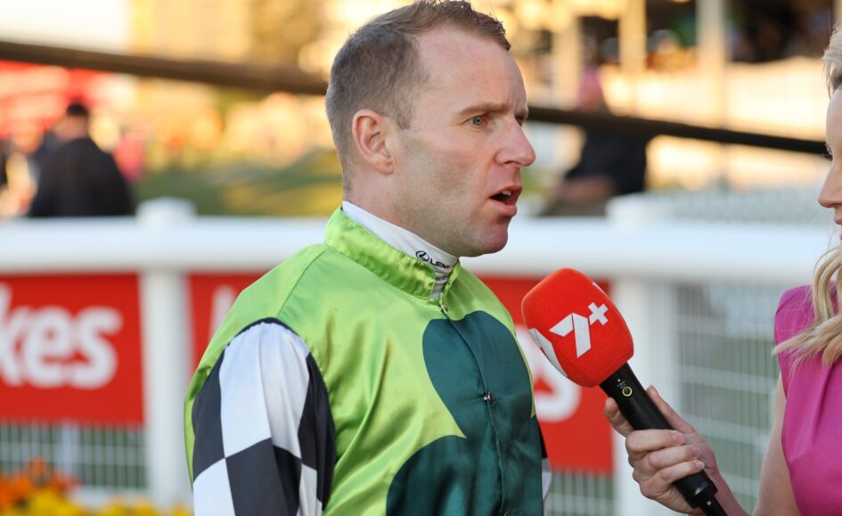 Man in a green racing suit being interviewed by a reporter with a red microphone outdoors on a sunny day.