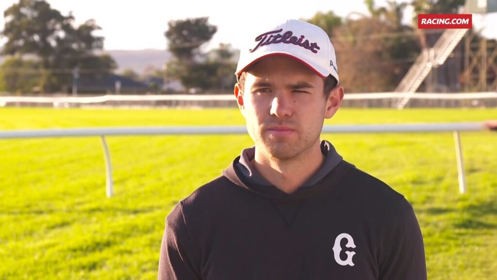 Portrait of a man in a Titleist cap and black hoodie on a sunny racecourse field with rails in the background.