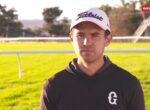 Portrait of a man in a Titleist cap and black hoodie on a sunny racecourse field with rails in the background.