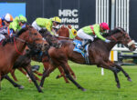 Jockeys in colorful silks race side by side on a grassy turf, leaning forward in a high-speed horse race image.