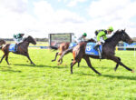 Jockeys ride racing horses on a grassy track under a blue sky with a large screen in the background.