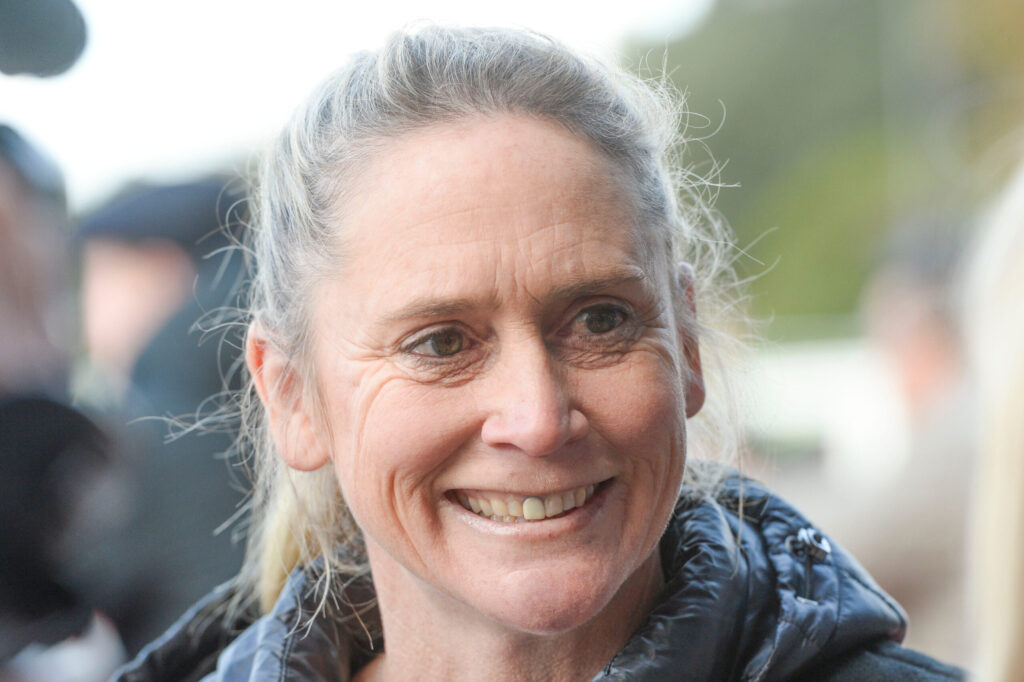 Close-up of a smiling older woman with gray hair outdoors, wearing a dark jacket.