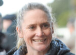 Close-up of a smiling older woman with gray hair outdoors, wearing a dark jacket.