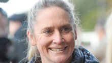 Close-up of a smiling older woman with gray hair outdoors, wearing a dark jacket.