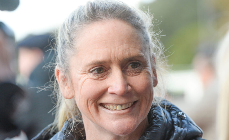Close-up of a smiling older woman with gray hair outdoors, wearing a dark jacket.