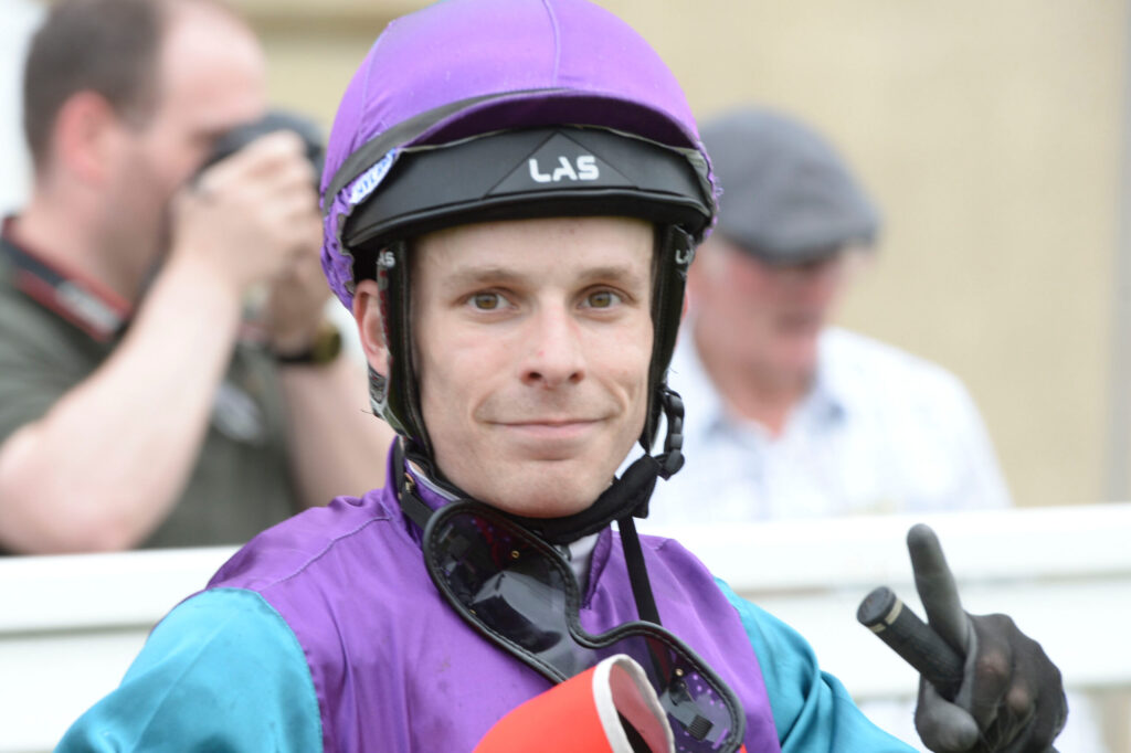 Close-up of a man wearing a purple helmet and purple-blue racing suit, looking at the camera with blurred people in the background.