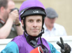 Close-up of a man wearing a purple helmet and purple-blue racing suit, looking at the camera with blurred people in the background.