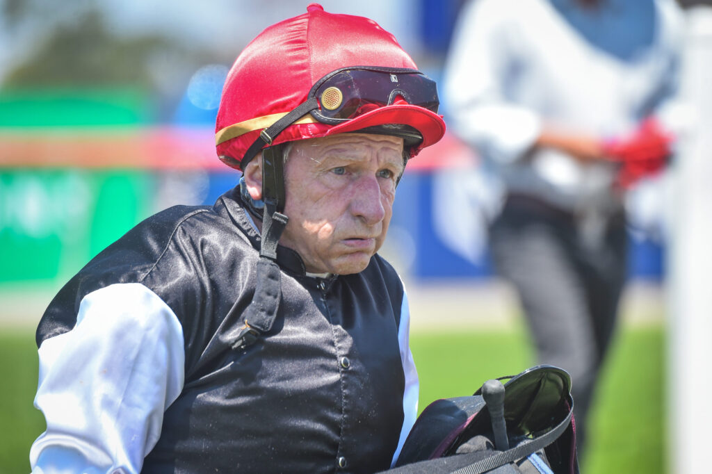 Older man wearing a red protective helmet and goggles, dressed in a black vest and white sleeves outdoors.
