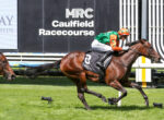 Jockey in orange and green silks riding a racehorse past the Caulfield Racecourse sign at a finish-style gate.