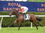 Jockey in red cap and white-and-red silks on a galloping bay horse during a race, with Royal Randwick signs in the background.