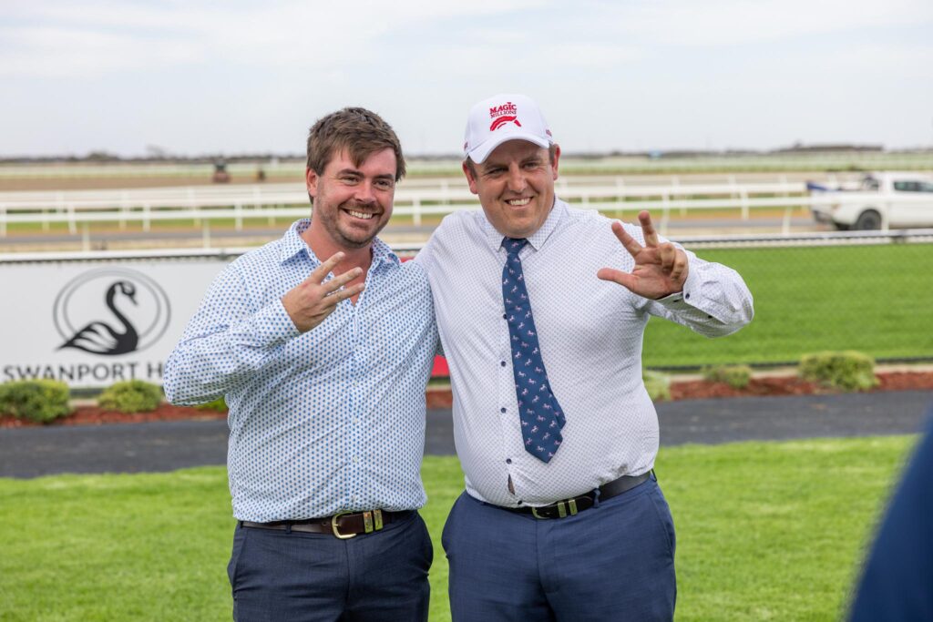 Two men in button-down shirts smile and pose with peace signs at a racecourse.