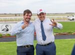 Two men in button-down shirts smile and pose with peace signs at a racecourse.