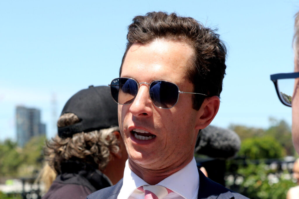 Close-up of a man in a suit and pink tie wearing aviator sunglasses outdoors on a sunny day, smiling with a blue sky behind him.