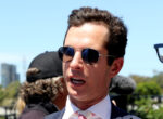 Close-up of a man in a suit and pink tie wearing aviator sunglasses outdoors on a sunny day, smiling with a blue sky behind him.