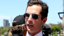 Close-up of a man in a suit and pink tie wearing aviator sunglasses outdoors on a sunny day, smiling with a blue sky behind him.