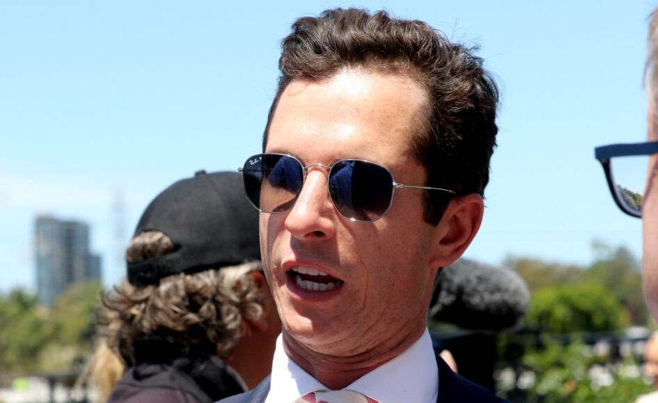 Close-up of a man in a suit and pink tie wearing aviator sunglasses outdoors on a sunny day, smiling with a blue sky behind him.