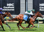 Two jockeys race on thoroughbreds at Caulfield Racecourse, sprinting neck-and-neck on a sunny turf track with the MRC Caulfield signage in the background.