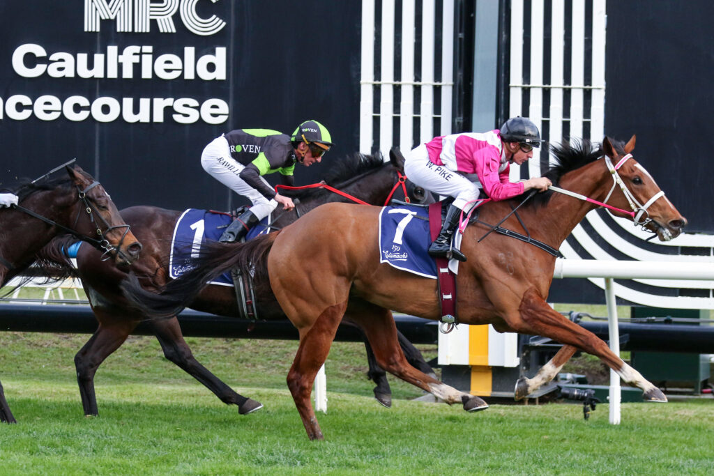 Horse race in progress with two jockeys leaning forward on sprinting horses, pink and green racing silks, blue and white saddlecloths, near a black-and-white grandstand backdrop.