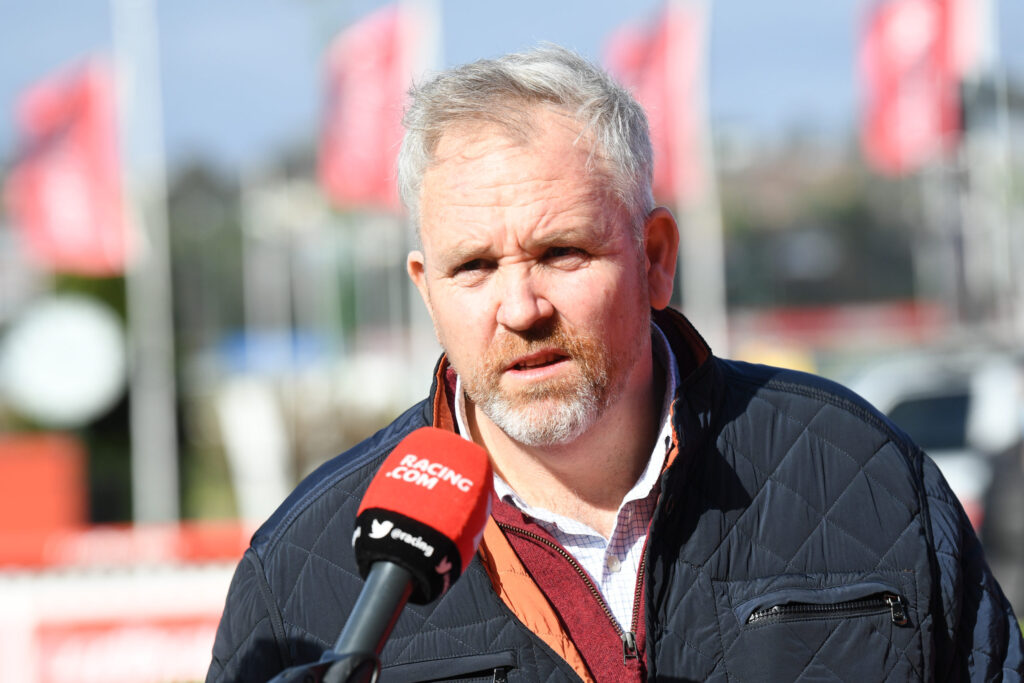Man being interviewed outdoors with a red Racing.com microphone, wearing a dark quilted jacket and white check shirt.