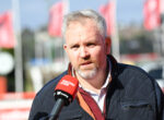 Man being interviewed outdoors with a red Racing.com microphone, wearing a dark quilted jacket and white check shirt.