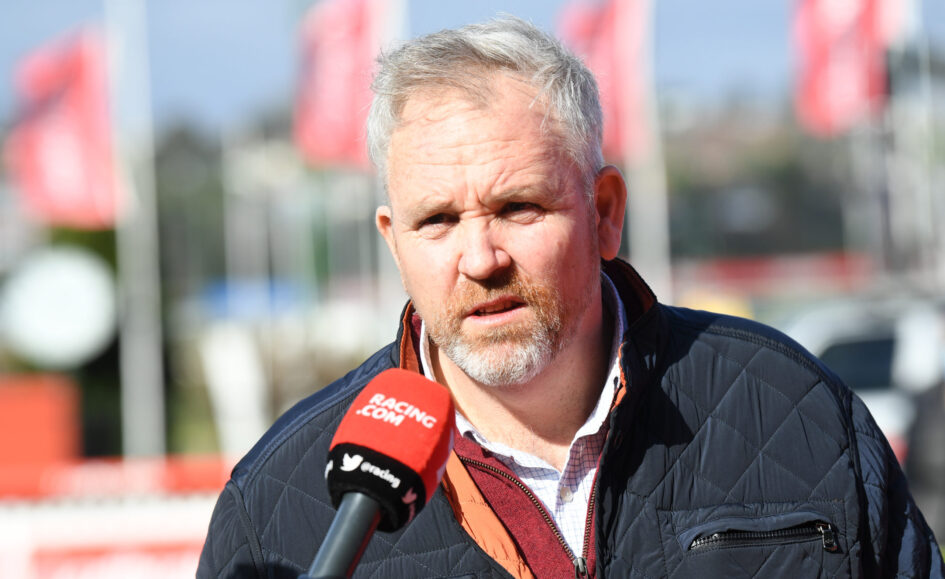 Man being interviewed outdoors with a red Racing.com microphone, wearing a dark quilted jacket and white check shirt.