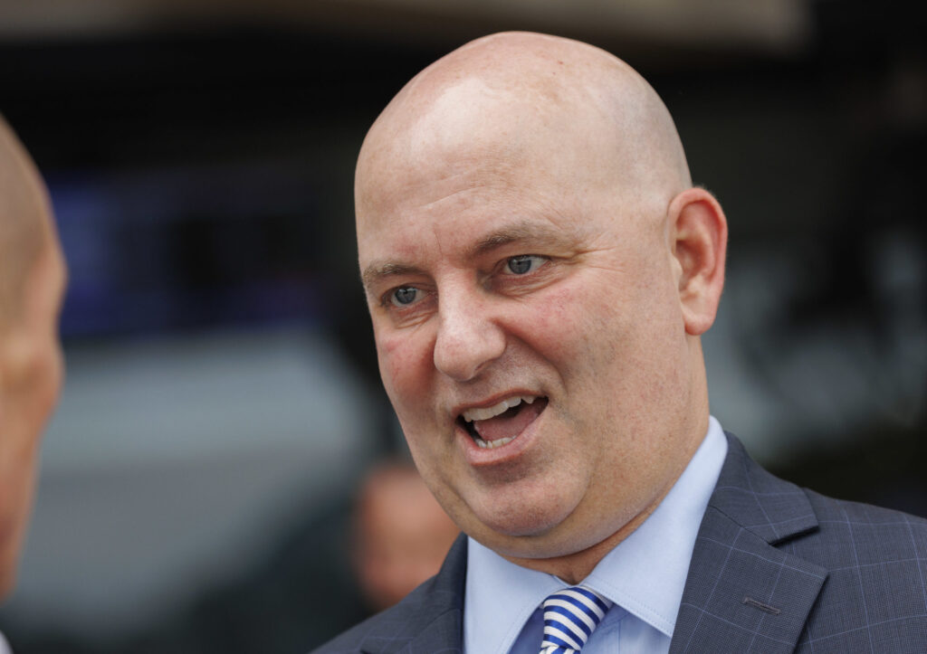 Close-up of a bald man in a blue suit and striped tie, mouth open as if speaking