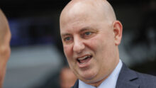 Close-up of a bald man in a blue suit and striped tie, mouth open as if speaking