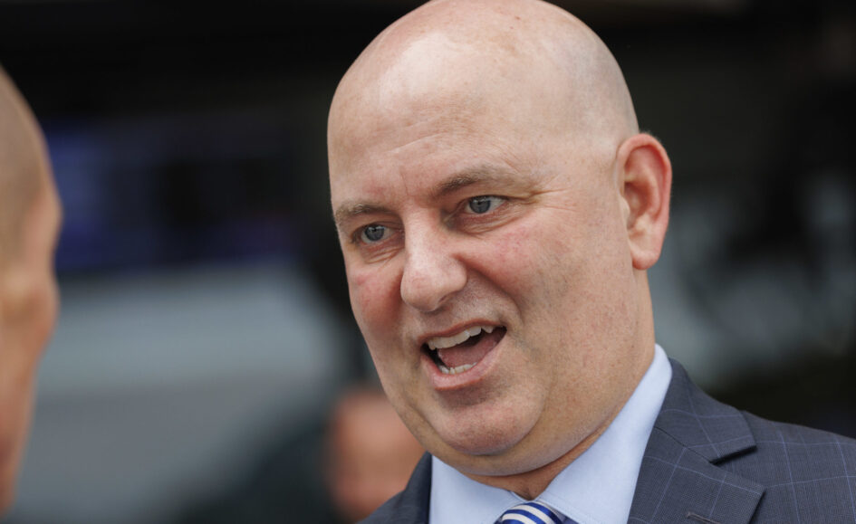 Close-up of a bald man in a blue suit and striped tie, mouth open as if speaking