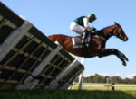 Horse and rider in green attire leap over a wooden obstacle in a grassy field under a clear blue sky.