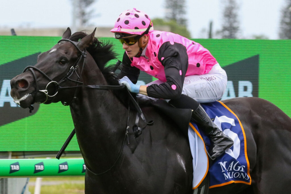 Jockey wearing a pink polka-dot helmet and matching pink shirt rides a dark horse during a race, with a green track barrier in the background.