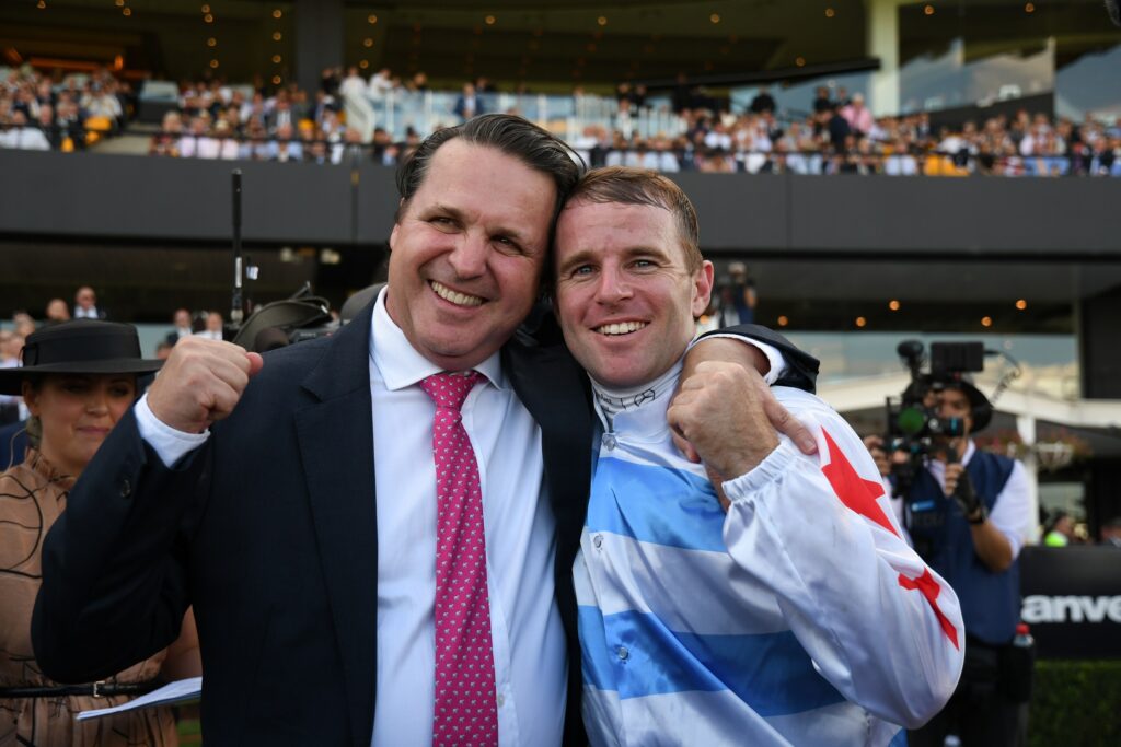 Two men smile and hug in celebration at a stadium, one in a dark suit and pink tie, the other in a blue-and-white flag jacket with a raised fist.
