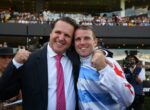 Two men smile and hug in celebration at a stadium, one in a dark suit and pink tie, the other in a blue-and-white flag jacket with a raised fist.
