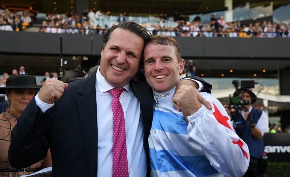 Two men smile and hug in celebration at a stadium, one in a dark suit and pink tie, the other in a blue-and-white flag jacket with a raised fist.