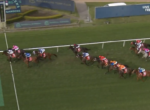 Horse race in progress with a pack of jockeys close together near the finish line on a grassy track.