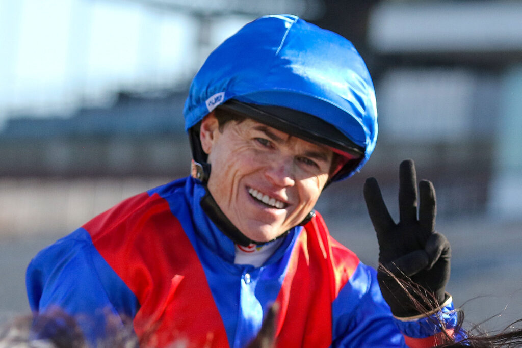 Smiling person wearing a blue helmet and red-and-blue jacket, raising a gloved hand toward the camera outdoors