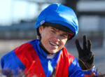 Smiling person wearing a blue helmet and red-and-blue jacket, raising a gloved hand toward the camera outdoors