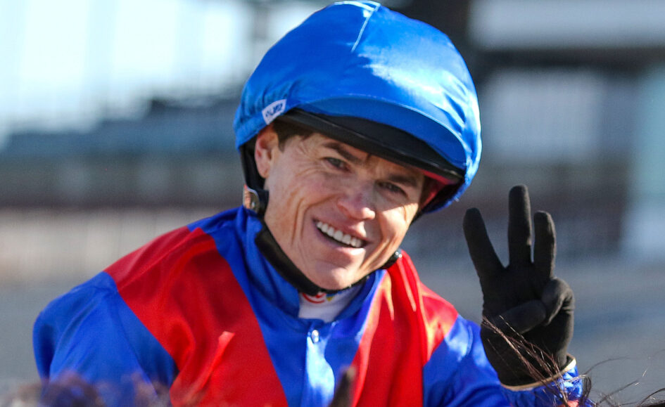 Smiling person wearing a blue helmet and red-and-blue jacket, raising a gloved hand toward the camera outdoors