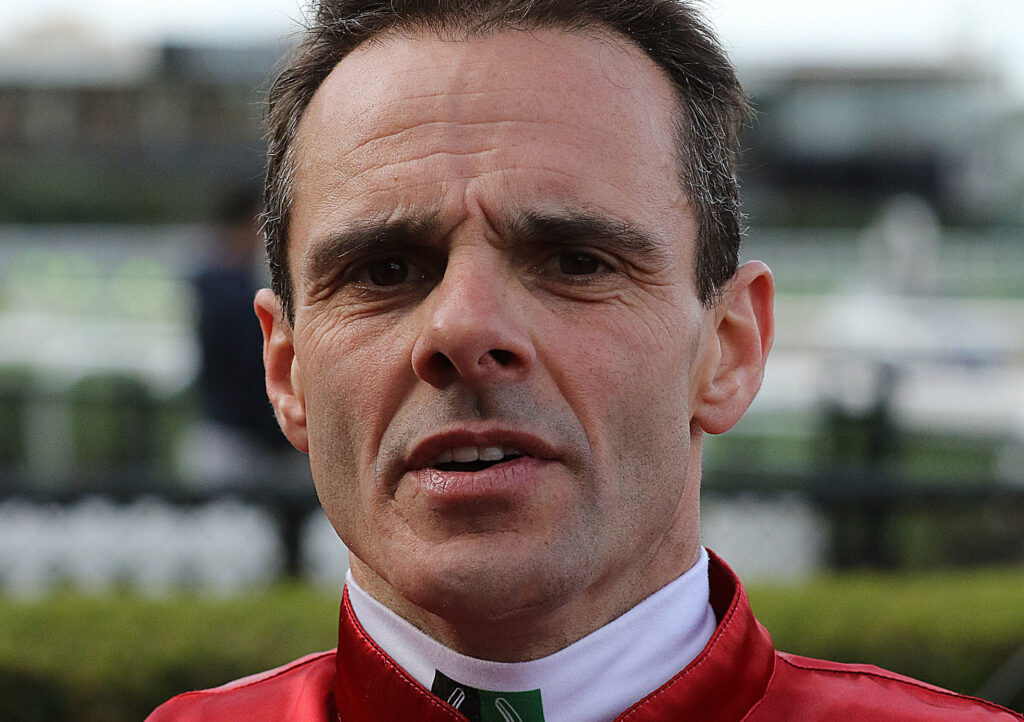 Close-up portrait of a man in a red racing jacket outdoors, focusing on his face from the nose up.