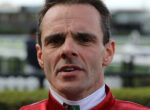 Close-up portrait of a man in a red racing jacket outdoors, focusing on his face from the nose up.