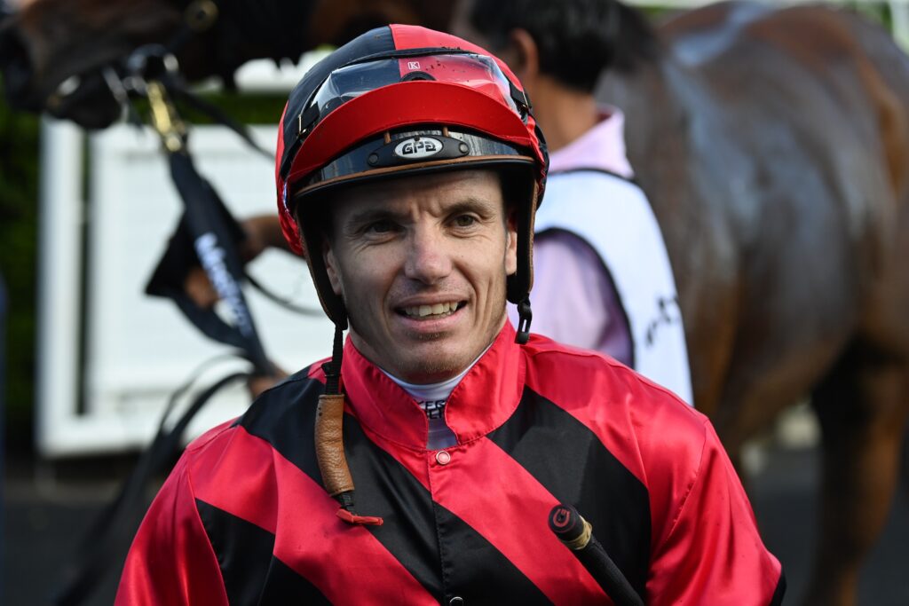 Close-up of a man in a red and black racing jacket and helmet, smiling, with a horse blurred in the background.