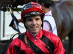 Close-up of a man in a red and black racing jacket and helmet, smiling, with a horse blurred in the background.