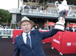 Man in a navy suit and straw hat lifts a large silver trophy at an outdoor event, with spectators behind him.