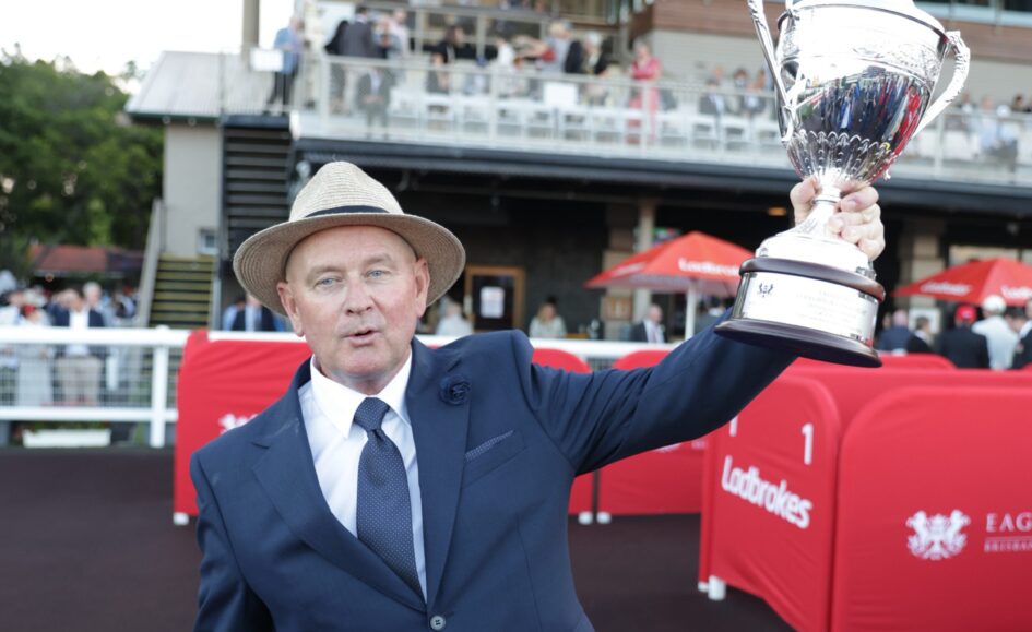 Man in a navy suit and straw hat lifts a large silver trophy at an outdoor event, with spectators behind him.