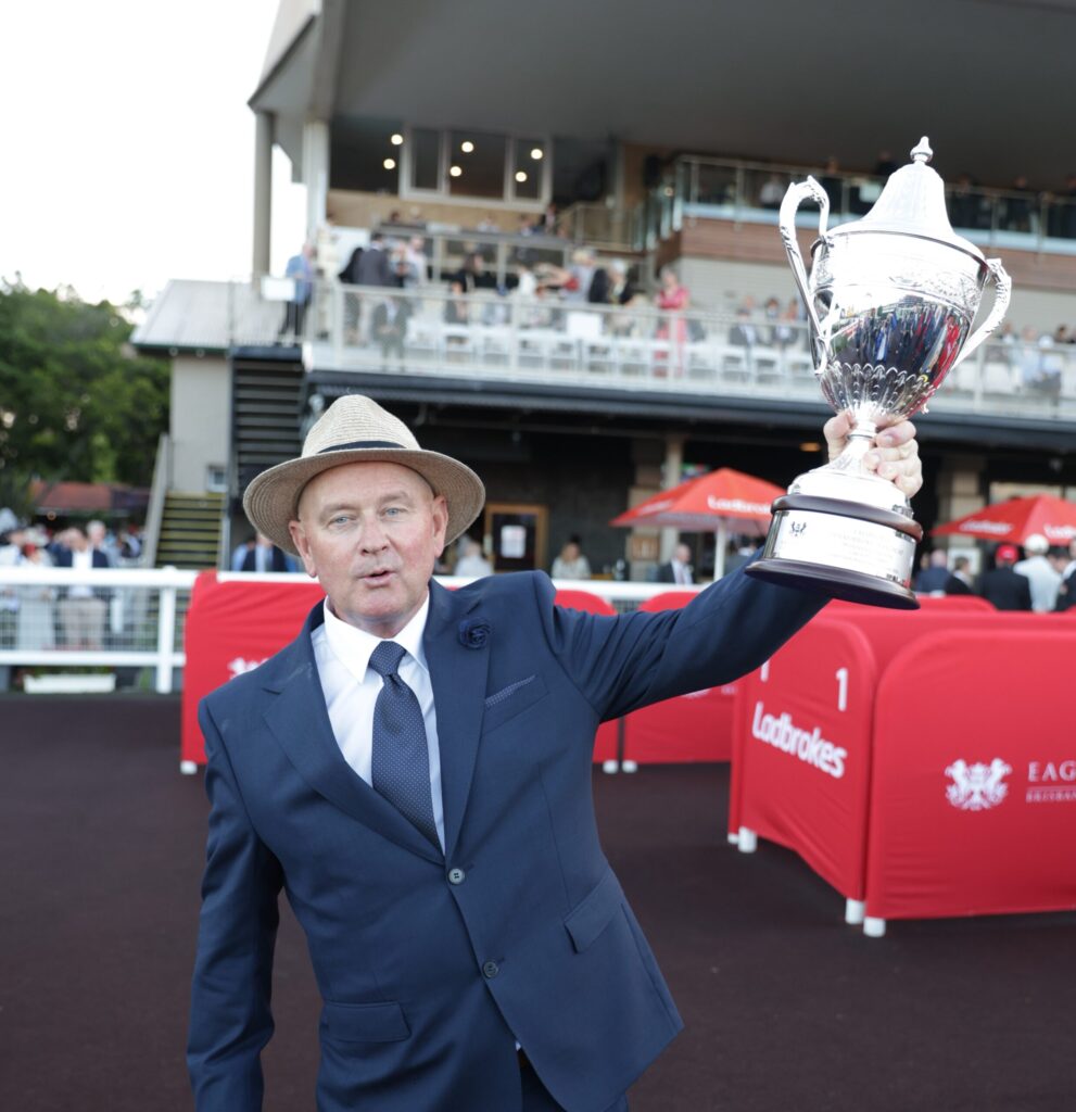 Man in a navy suit and straw hat lifts a large silver trophy at an outdoor event, with spectators behind him.